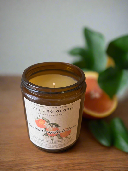 A brown jar candle with a label that reads 'Orange Grapefruit Grove', with a lit flame, alongside a slice of orange and green leaves in the background.