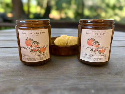 Two jars of SOLI DEO GLORIA candles, one labeled 'Orange Grapefruit Grove', with a small wooden bowl of wax tarts in front of them, set on a wooden surface with foliage in the background.