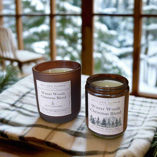Two candles labeled 'Winter Woods Christmas Blend' on a wooden surface with a snowy window background.