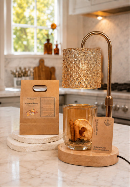 A candle set displayed on a marble kitchen counter, featuring a clear glass jar candle with visible cinnamon sticks and dried botanicals inside, a wooden base, a textured gold-toned lamp above it, a kraft paper bag package labeled Cinnamon Harvest, and a small product tag. The background shows a softly blurred kitchen interior with a window and cabinetry.