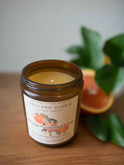 A brown jar candle with a label that reads 'Orange Grapefruit Grove', with a lit flame, alongside a slice of orange and green leaves in the background.