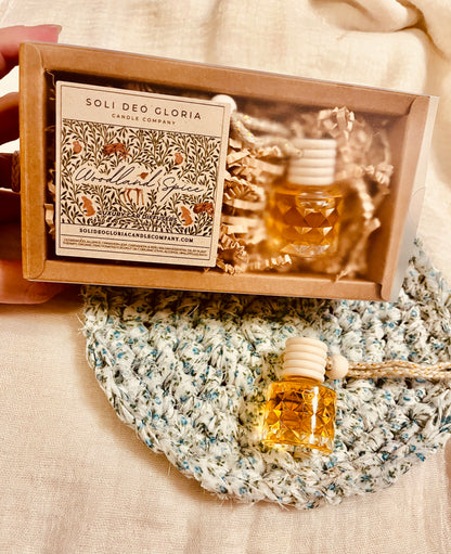 A hand holds an open kraft gift box containing a Soli Deo Gloria candle and a small amber glass diffuser bottle with a white wooden stopper, surrounded by shredded paper. A matching small diffuser bottle rests on a blue-and-white knit blanket on a beige fabric surface.