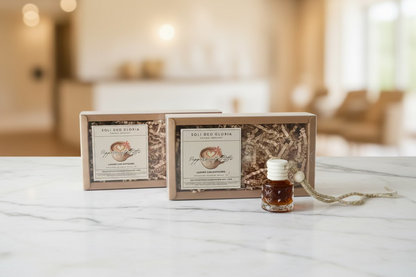 Two rectangular gift boxes labeled 'SOLI DEO GLORIA' filled with shredded brown paper, placed on a wooden table, with a small amber glass bottle and a silver beaded necklace in front of them.