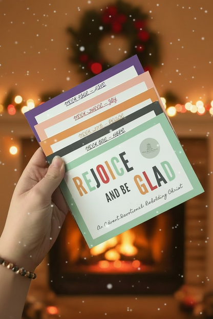 A hand holds a fanned stack of colorful Advent devotional cards in a busy indoor setting. The front card reads 'REJOICE AND BE GLAD' with pastel purple, peach, orange, gray, and mint green borders and text.