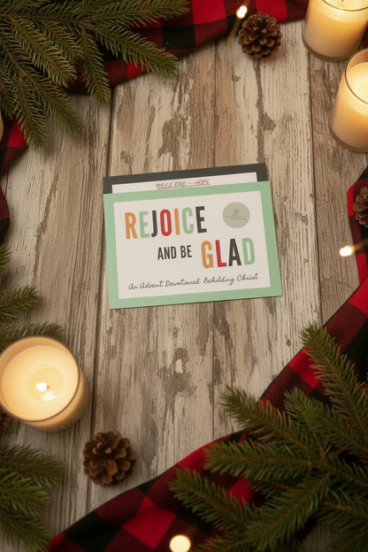 A hand holds a stack of colorful Advent devotional cards in a busy indoor setting. The front card reads 'REJOICE AND BE GLAD' with smaller text 'An Advent Devotional Believable Christ' and a small circular logo.